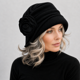 A woman wearing a black slouchy cloche hat with a large fabric flower accent on the side, paired with a black turtleneck top against a neutral gray background.