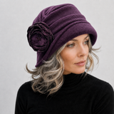 A woman wearing a deep purple woolen cloche hat with a large fabric flower embellishment on the side, paired with a black turtleneck sweater against a light gray background.